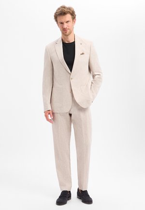 Man with light beige suit, black shirt, and black loafers stands with one hand in pocket against a white background.