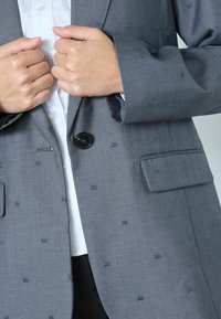 Blazer gris con patrón texturizado, cierre de un solo botón negro, dos bolsillos frontales, corte entallado y puño de camisa visible por debajo.