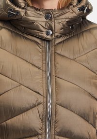 Veste matelassée légère marron clair avec une fermeture éclair centrale argentée, un col haut et des cordons noirs sur une capuche.