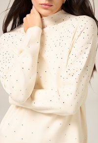 Kremowy sweter z długimi rękawami, zdobiony różnorodnymi srebrnymi kwadratowymi aplikacjami, z wysokim dekoltem zapewniającym dopasowany wygląd.