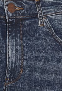 Tmavě modré džíny s hladkou texturou, kontrastním prošíváním a měděným knoflíkem v pase. Přítomné jsou vybledlé oblasti a jemné detaily vrásek.