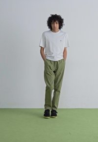 White short-sleeve t-shirt, green relaxed-fit pants, and black sneakers with a yellow sole, standing against a light background.