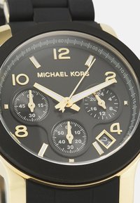 Reloj con correa de goma negra y esfera dorada y negra. Presenta tres subesferas, marcadores dorados y una pantalla de fecha en la posición de las 4.