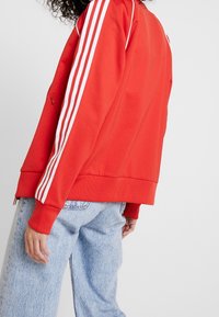 Červená sportovní bunda s bílým třídílným detailem na rukávech, žebrovanými manžetami a spodním lemem, kombinovaná se světle modrými džíny.