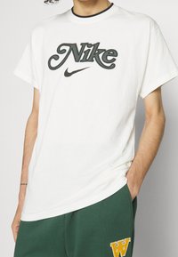 Vit bomull t-shirt med en klassisk Nike-logotyp i mörkgrön och svart, med rund halsringning och korta ärmar.