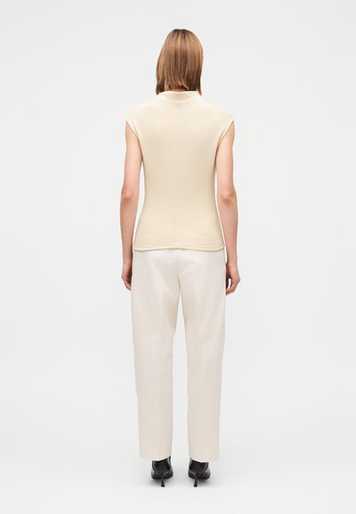 Figursydd beige stickad topp med korta ärmar, kombinerad med lösa, ljusa byxor och svarta skor med klack, sedd bakifrån.