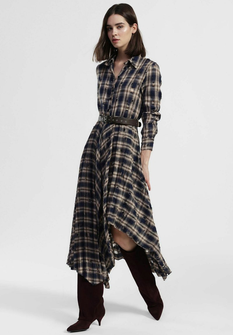 Vestito camicia a quadri scuri con orlo asimmetrico, maniche lunghe e cintura marrone. Presenta una chiusura con bottoni sul davanti e una texture del tessuto morbida.