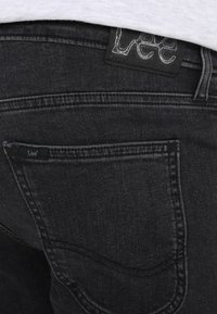 Jeans de mezclilla negros con detalle de bolsillo trasero bordado, que presentan una etiqueta de cuero con "Lee" grabado en la cinturilla. Textura suave, corte ajustado.