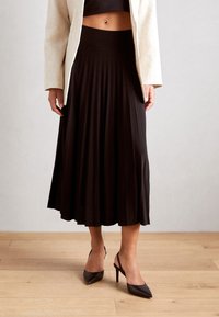 Gonna nera plissettata con una vita alta, abbinata a un crop top nero aderente e a un blazer beige, indossata con scarpe con tacco alto nere.