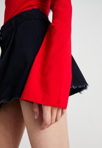 Haut rouge à manches longues avec des manches évasées, associé à une jupe noire avec un ourlet effiloché. Accent sur la texture du tissu et le contraste des couleurs.