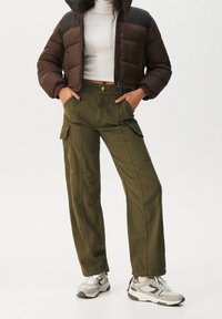 Kvinna står självsäkert i en brun puffjacka och vit turtleneck, iklädd olivgrön cargobyxor och grå sneakers, med händerna i fickorna.