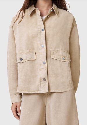 Mujer con chaqueta beige con botones en la parte delantera con dos bolsillos delanteros con solapa y pantalones holgados a juego.