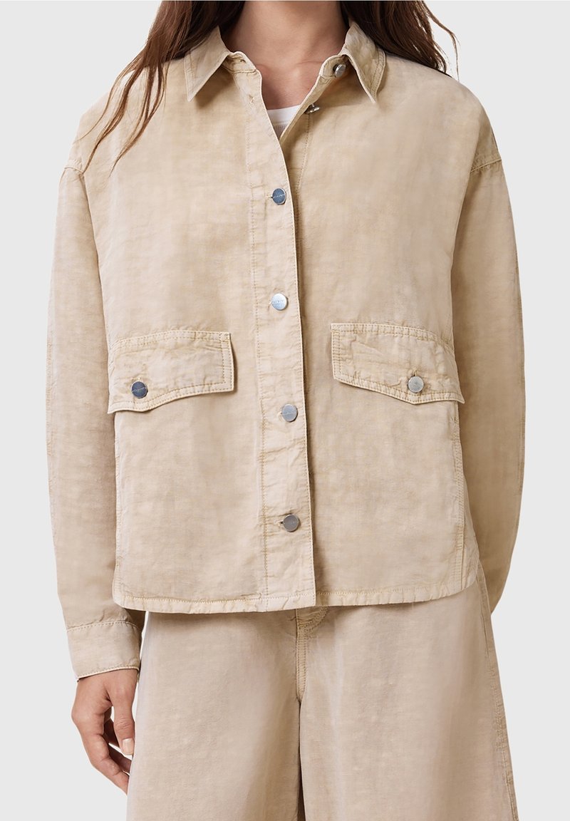 Femme portant une veste beige à boutons avec deux poches à rabat à l'avant et un pantalon ample assorti.