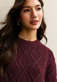 Maglione a maglia color bordeaux con un motivo a diamante testurizzato, scollo a costine e vestibilità rilassata. Il modello indossa orecchini a cerchio e ha i capelli lunghi.