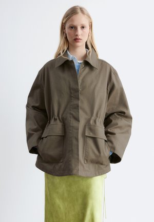 Jeune femme portant une veste olive oversize avec de grandes poches avant et une jupe vert clair, debout devant un fond blanc uni.