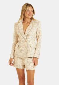 Blazer en tweed beige avec un design à double boutonnage, revers crantés, deux poches avant et un short assorti, affichant une finition texturée.