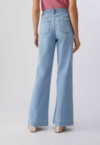 Femme portant un jean ample taille haute bleu clair et des talons en maille beige, debout dos à un fond clair uni.