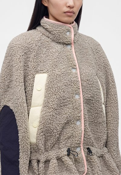 Γυναίκα που φοράει γκρι ριγωτό fleece μπουφάν με κουμπιά πατιλέτας, ροζ περιθώριο, κρεμ καπιτονέ τσέπες και μαύρα επιθέματα στους αγκώνες.