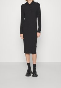 Schwarzes geripptes Kleid mit einem kragenförmigen Ausschnitt, langen Ärmeln und Knopfleiste. Getragen mit schwarzen Socken und Schuhen, knöchellang.