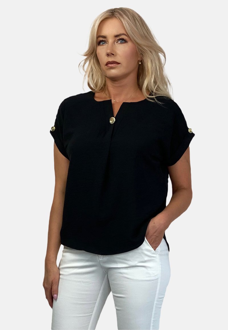 Czarna bluzka krótki rękaw z dekoltem w serek, z nadrukowanymi złotymi guzikami na rękawach i luźnym krojem, połączona z białymi spodniami. Gładka tekstura.