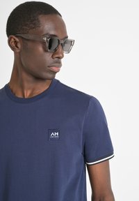 Camiseta de cuello redondo navy con un parche de logo, con mangas cortas y acentos en rayas blancas. Modelo usando gafas de sol transparentes.
