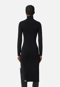 Robe noire en tricot avec un col montant, des manches longues et une fente sur le côté ; présente un détail logo près de l'ourlet et une coupe ajustée.