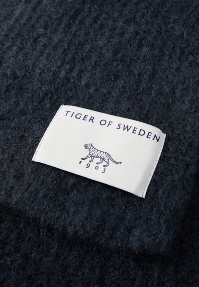 Hvit stoffetikett med "Tiger of Sweden" og tigerlogo, sydd på mørkeblå teksturert strikket stoff.