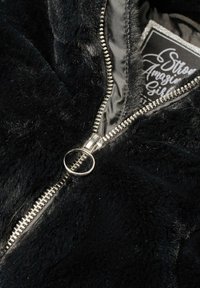 Gros plan d'une veste noire en fourrure avec un tirette de fermeture éclair circulaire argentée partiellement ouvert, montrant une étiquette avec un texte blanc décoratif à l'intérieur.