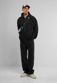 Black zip-up hoodie with front pockets, matching black pants, and cream sneakers. The model wears a black-and-white cap and a crossbody bag.