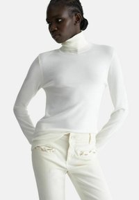 Maglione bianco a collo alto abbinato a jeans crema, caratterizzato da un tessuto texturizzato, una vestibilità slim e dettagli decorativi sulle tasche.