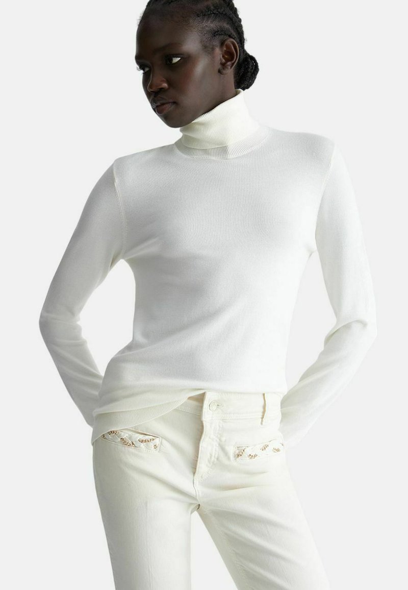 Maglione bianco a collo alto abbinato a jeans crema, caratterizzato da un tessuto texturizzato, una vestibilità slim e dettagli decorativi sulle tasche.