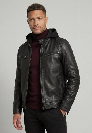 Mand iført sort læderjakke med hætte over mørkrød rullekravesweater, stående mod en ensfarvet grå baggrund.