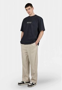 Tricou mare de culoare albastru închis, cu un grafic pe față, pantaloni bej cu dungi laterale, adidași alb-negru. Material neted, croială confortabilă.
