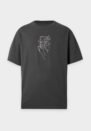 Mann trägt ein schwarzes T-Shirt mit Grafik, helle Jeans-Knielange Shorts und schwarze Sandalen und steht vor einem einfarbigen weißen Hintergrund.