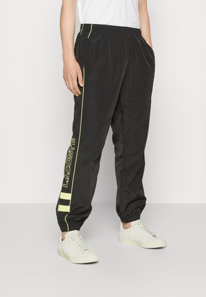 Sorte træningsbukser med elastiske manchetter og gule sidelister med "Lacoste"-logoet, båret med hvide sneakers og en hvid skjorte.