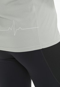 Lys grå træningsshirt med et grafik af en hjerterytme linje i kanten, parret med sorte teksturerede leggings med mesh-paneler.