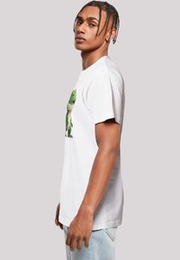 White cotton t-shirt featuring a colorful dinosaur graphic on the front, round neck, and short sleeves. Model poses sideways.