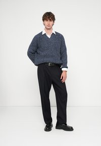 Camisola grossa de malha azul-marinho com decote em V, combinada com calças pretas de pernas largas e sapatos pretos grossos. Gola de camisa branca visível por baixo.