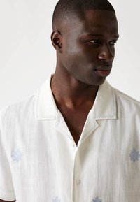 White short-sleeve shirt with blue embroidered patterns, a soft texture, and a button closure. Features a camp collar and relaxed fit.
