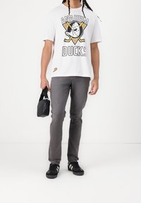 Vit t-shirt med "ANAHEIM DUCKS"-logotyp och grafik, grå jeans, svarta sneakers med vita ränder, håller en liten svart väska.