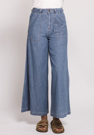 Wide-leg blue denim pants with front patch pockets worn with beige studded sandals.