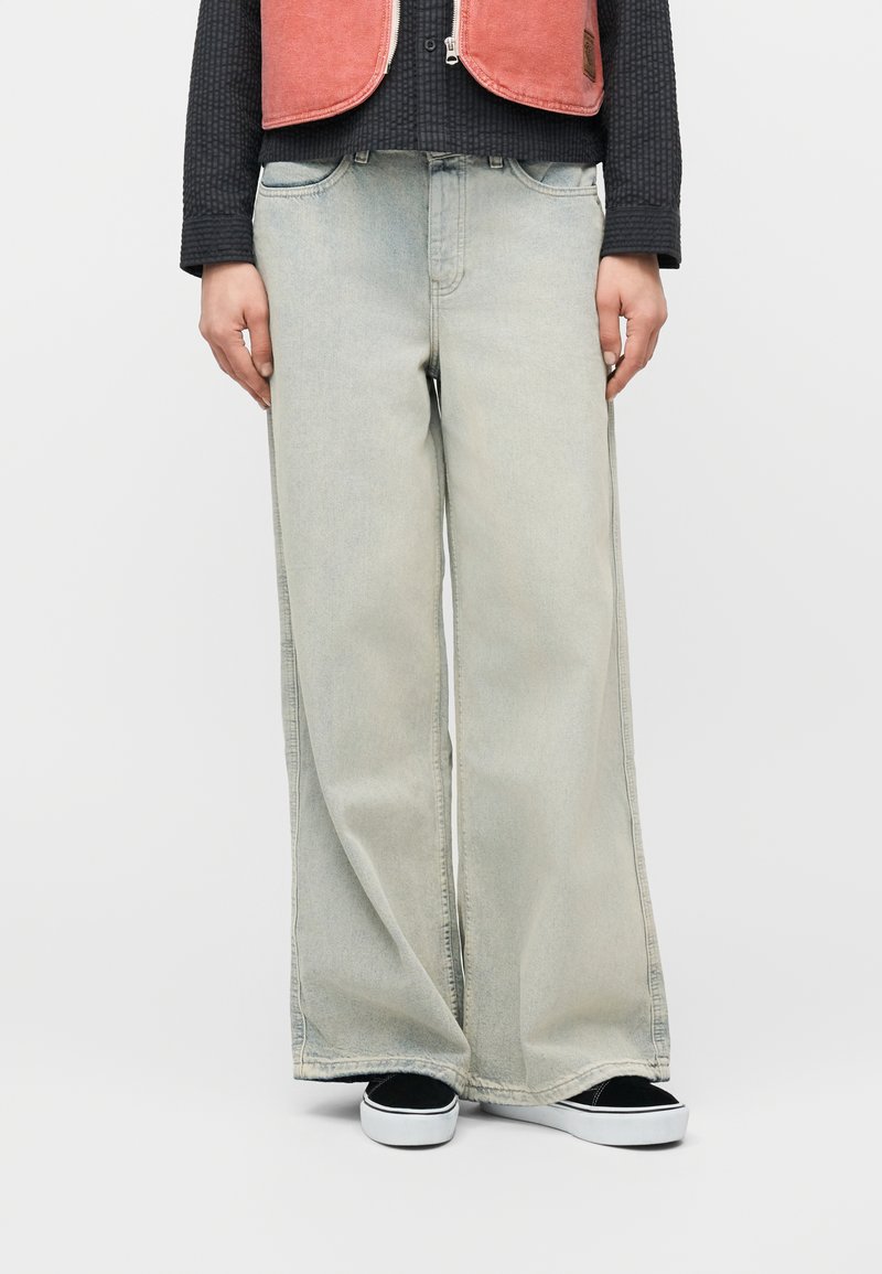 Personne portant un jean gris clair à jambes larges, un haut noir à manches longues, un gilet zippé corail, et des baskets noires à enfiler avec des semelles blanches.