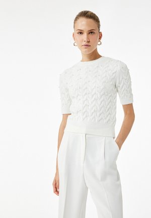 Weißer Strickpullover mit strukturiertem Netzdesign, kurzen Ärmeln und Perlenakzenten. Kombiniert mit hochgeschnittenen weißen Hosen.