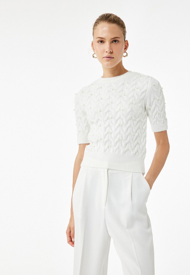 Valkoinen neulepaita, jossa on teksturoidut vyyhtimuotoilut, lyhyet hihat ja helmiakcentit. Yhdistetty korkeavyötäröisten valkoisten housujen kanssa.