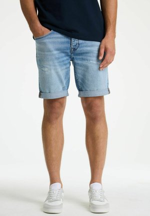 Man wearing light blue rolled denim shorts, white sneakers, and a dark t-shirt, standing with one hand in pocket.