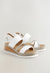 Des sandales à talon compensé blanches avec plusieurs larges sangles et des accents en argent. Elles présentent un bord inférieur texturé et une semelle rembourrée pour le confort.