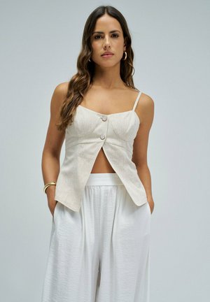 Femme aux longs cheveux ondulés portant un haut beige sans manches boutonné et un pantalon blanc ample, debout sur un fond clair uni, les mains dans les poches.