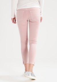 Persoon in lichtroze slim-fit broek met achterzakken en zilveren instappers, staand tegen een witte achtergrond.