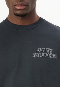 Tumši jūras zila kokvilnas svītra ar apaļu kakla izgriezumu un baltu izšūtu "OBEY STUDIOS" logotipu kreisajā krūtī.