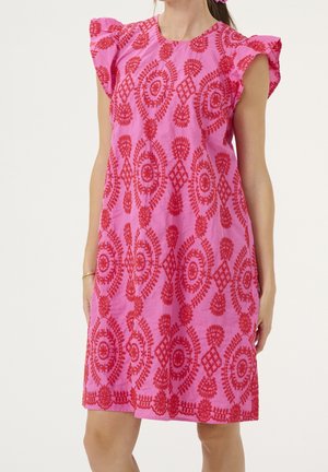 Różowa sukienka bez rękawów z falbanami na ramionach i czerwonym, misternym kwiatowym haftem, noszona przez stojącą kobietę na białym tle.